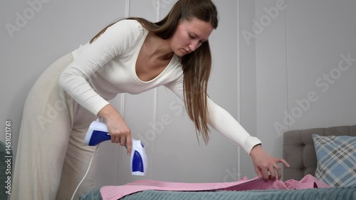 Housewife doing routine work Carefully ironing clothes on the bed in the home bedroom with a handheld steamer, demonstrating daily household chores and home care