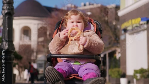 A child with curly hair happily eats a pretzel while sitting in a stroller in a bustling outdoor area The sun shines creating a cheerful atmosphere
