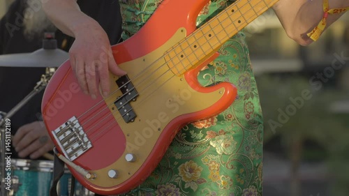 This video shows a close-up view of a woman strumming an orange bass guitar while wearing a tropical green retro dress during an outdoor performance. 