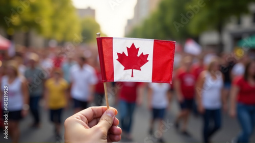 A close-up of a hand holding a small Canadian flag, with a blurred background of a Canada Day celebration.