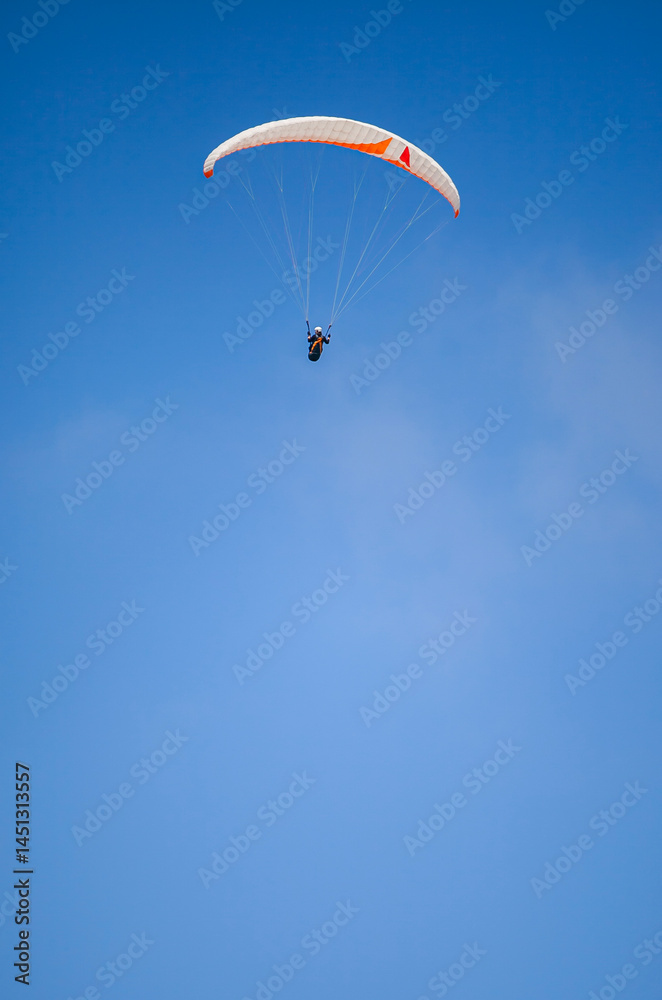 A paragliding athlete is floating in the air showing his skills in controlling a paraglider against a backdrop of bright blue sky.