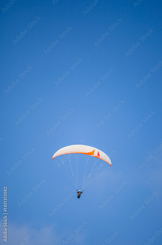A paragliding athlete is floating in the air showing his skills in controlling a paraglider against a backdrop of bright blue sky.