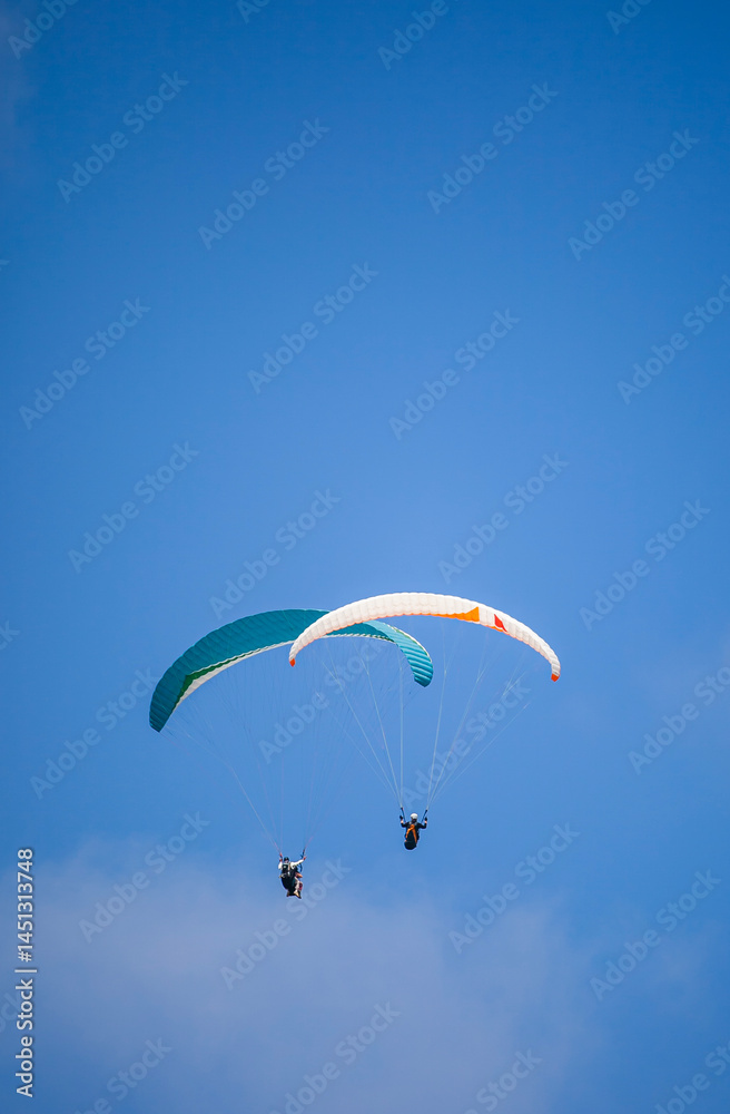 A pair of paragliding athletes are floating in the air showing their skills in controlling their paraglider against a backdrop of bright blue sky.