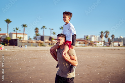 Father carrying son on shoulders walking on beach during summer vacation