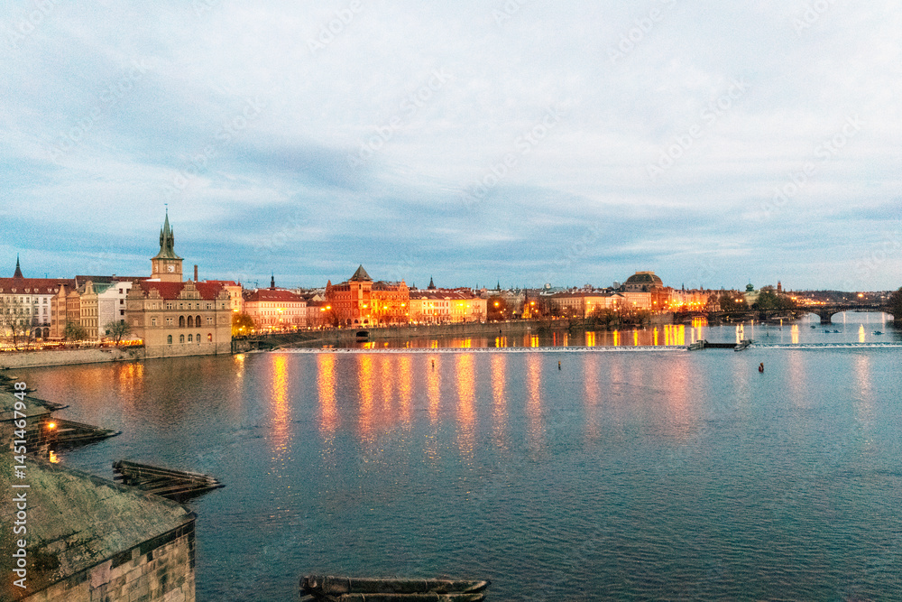 Naklejka premium A cityscape by the riverbank at dusk. The many lights of the city's buildings reflect in the river water, creating a warm and glowing effect.