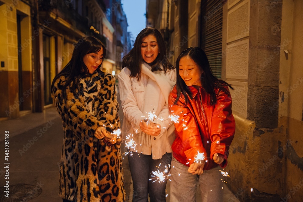 Fototapeta premium A dark-haired Caucasian woman in her early 20s, a European adult woman, and an Asian young woman wearing in warm jackets holding lit sparklers and smiling in a narrow street in the evening.