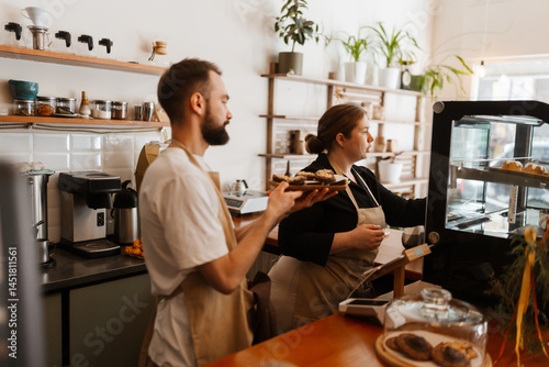 Obraz na płótnie Side view of a dark-haired adult male barista in a white t-shirt holding desserts while an attentive female European owner in her early 30s puts them in the fridge at the coffee shop. w salonie