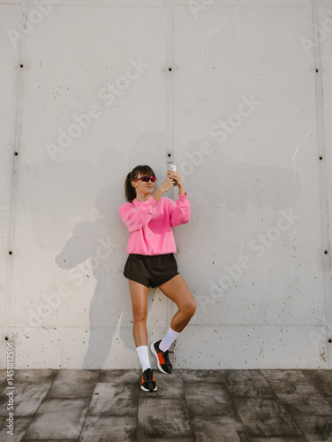 A European sportswoman in her 30s with dark hair tied in a ponytail dressed in sunglasses, a pink sweatshirt, shorts, and sneakers taking a selfie while standing in front of a white wall in the street