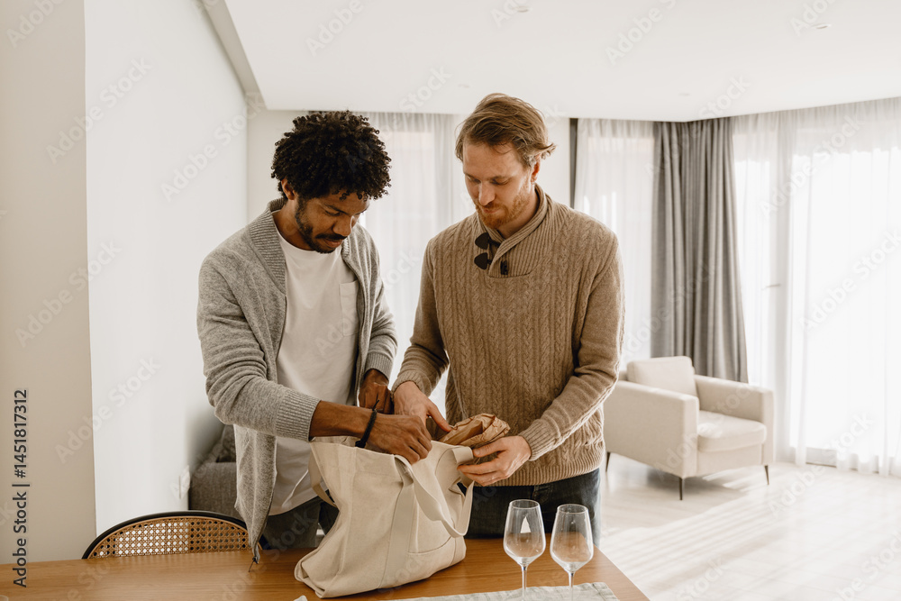 Fototapeta premium A multiethnic gay couple in their 30s dressed in sweaters and jeans standing at a table and packing a bag together in a cozy room with white walls, big windows, and a white armchair in the background