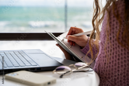 Woman writing ideas in notebook, sitting at table, taking notes, brainstorming, journal and pen for planning. Female write in copybook in cafe. Work, study. Cropped close-up