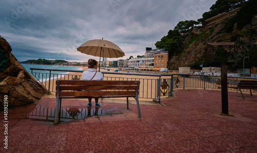 Pretty woma with an umbrella sits on the a bench with city beach and stormy clouds in the sky background. Lloret de Mar, Girona region, Spain.