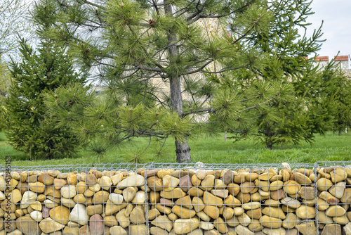 Stone fence reinforced with strong galvanized metal mesh, made of large smooth pebbles against backdrop of summer park. Copy space for text.