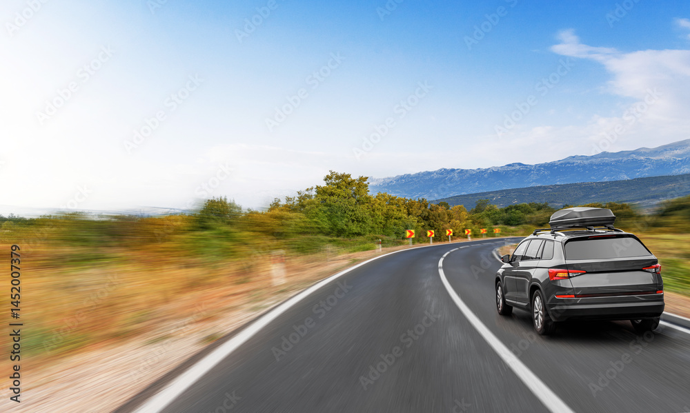 Fototapeta premium A modern SUV with a rooftop cargo box drives along a winding country road surrounded by trees and mountains under a clear blue sky.