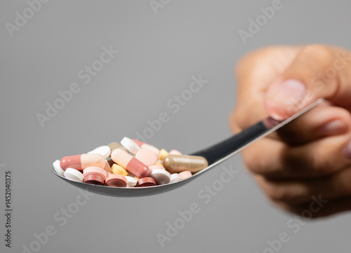 Conceptual image: A hand holding a spoon filled with multicolored pills and capsules, against a gray background.