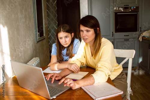 Mother and daughter using laptop sitting together in home kitchen. Mom helping kid with school homework. Education with modern technologies.