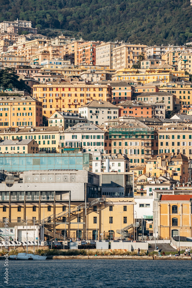 Obraz premium Genoa, Italy - September 22, 2024: Colorful hillside facades and layered historic buildings rise above the port waterfront with boats and greenery.