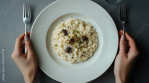 Flat lay image of creamy truffle risotto on a round white plate positioned on the left side of a cool grey background, two hands holding silver fork and knife, photo