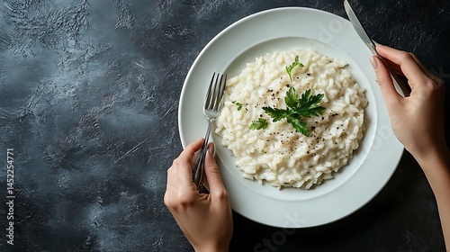 Flat lay image of creamy truffle risotto on a round white plate positioned on the left side of a cool grey background, two hands holding silver fork and knife, photo