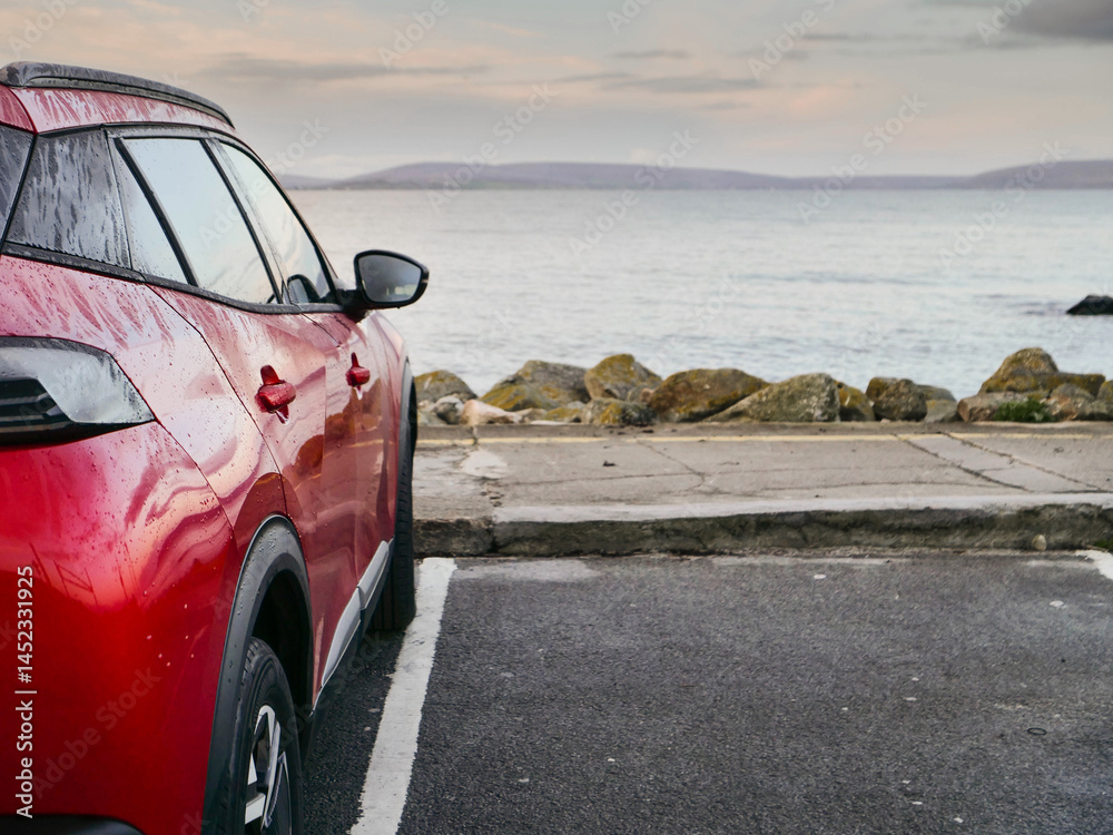 Red car parked in car park with stunning view on nature scenery with ocean and mountains and cloudy sky. Silverstrand area, Galway city, Ireland. Rent motor for a tourist trip concept.