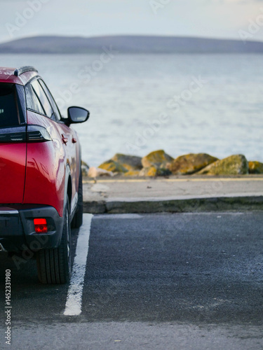 Obraz na płótnie Red car parked in car park with stunning view on nature scenery with ocean and mountains and cloudy sky. Silverstrand area, Galway city, Ireland. Rent motor for a tourist trip concept. w salonie