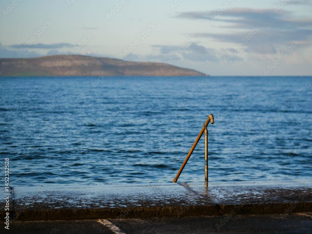 Metal hand rail and wet foot path in focus in foreground. Blue ocean surface of Galway bay and Burren mountains in the background. Calm and relaxing nature scene. Nobody. Travel and tourism.