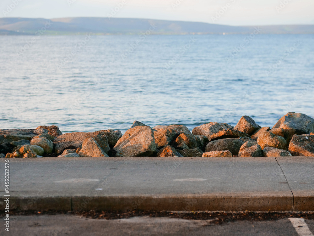 Foot path by big rocks and blue ocean surface and mountains in the background. Travel and tourism. Silverstrand, Galway city, Ireland. Nobody. Nature scenery and popular outdoor activity area.