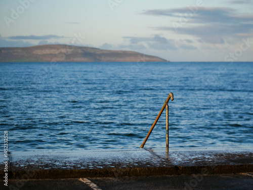 Obraz na płótnie Metal hand rail and wet foot path in focus in foreground. Blue ocean surface of Galway bay and Burren mountains in the background. Calm and relaxing nature scene. Nobody. Travel and tourism. w salonie