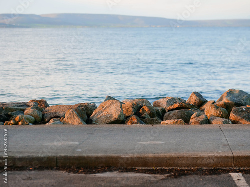 Obraz na płótnie Foot path by big rocks and blue ocean surface and mountains in the background. Travel and tourism. Silverstrand, Galway city, Ireland. Nobody. Nature scenery and popular outdoor activity area. w salonie