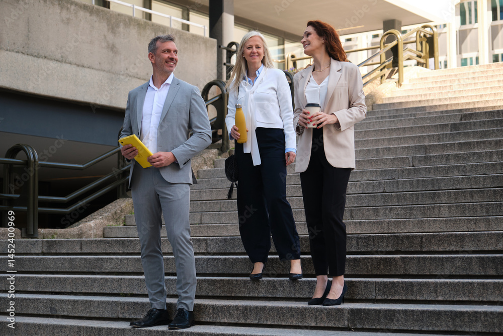 Business team walking down the stairs, holding tablet, reusable water bottle and takeaway coffee, talking and smiling