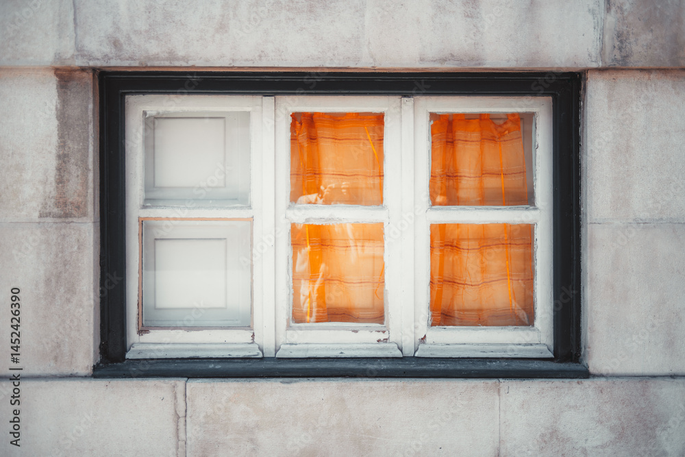 Obraz premium Close-up of an old wooden-framed window set in a stone wall, with vivid orange curtains partially drawn inside, creating a warm and cozy contrast against the weathered exterior