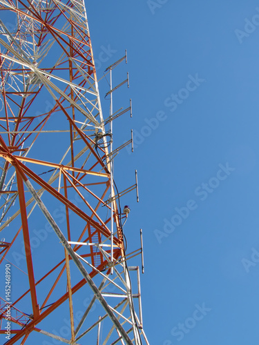 A tall orange and white self-supporting telecommunications tower with a 6 dipole array stands against a clear blue sky transmitting radio signals.