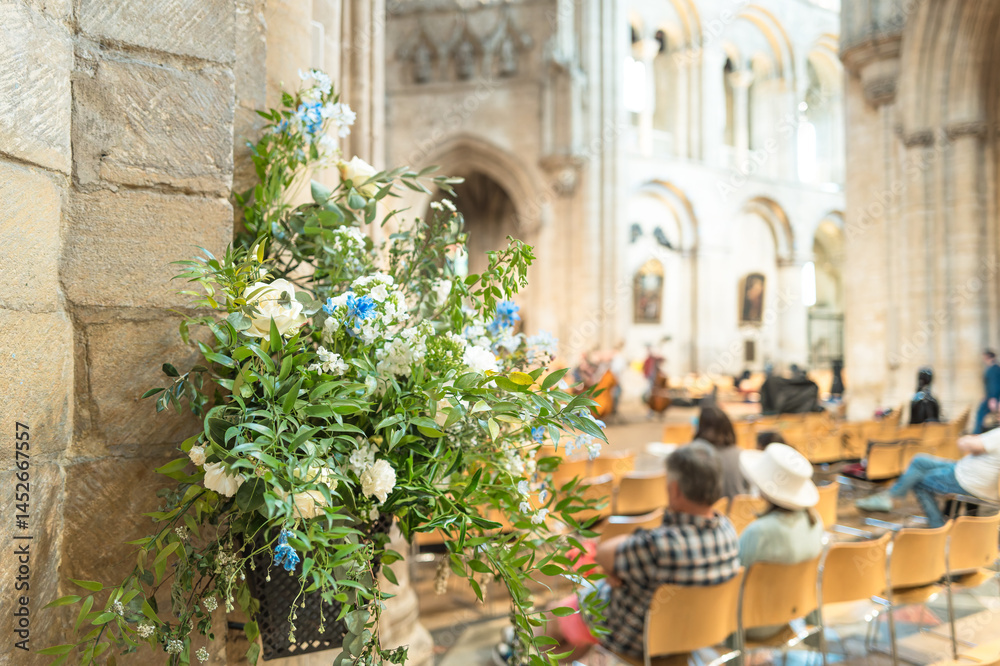 Shallow focus of an artificial flower arrangement seen on a pillar of a famous English cathedral. People can be sitting on chairs within the cathedral listing to a live orchestrator practice.