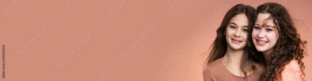 Banner with studio portrait of two beautiful smiling sisters in peach and beige shirts against peach background