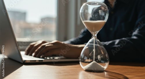 Person using laptop with hourglass on desk time management concept.