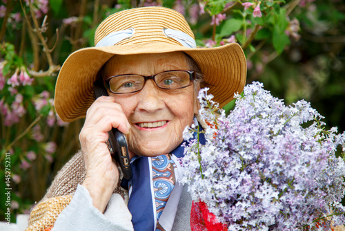 Portrait of an old woman with a mobile phone and a bouquet of lilacs. An elderly woman thanks on the phone for a bouquet of lilacs. The concept of boarding houses, old age, care. 