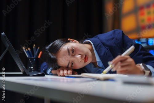 Young Asian manager falling asleep on desk while working late in office at night, suffering from chronic fatigue or overwork