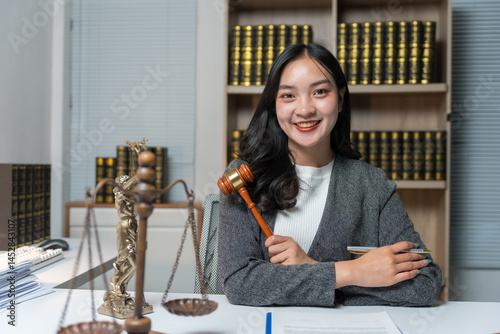 Asian female lawyer smiling and holding a gavel in her office with scales of justice and law books