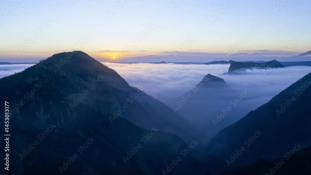 Mystical mountain range silhouetted against dawn sky with fog rolling through valleys. The view captures dramatic peaks rising above cloud cover as soft morning light gradually illuminates the horizon