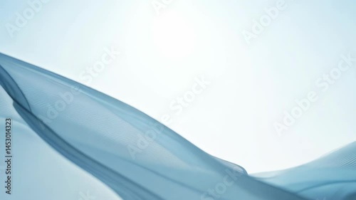 Flowing blue silk fabric gradually unfurling against white background. The translucent material creates wave patterns as it moves from left to right, displaying smooth curves and gentle movement.