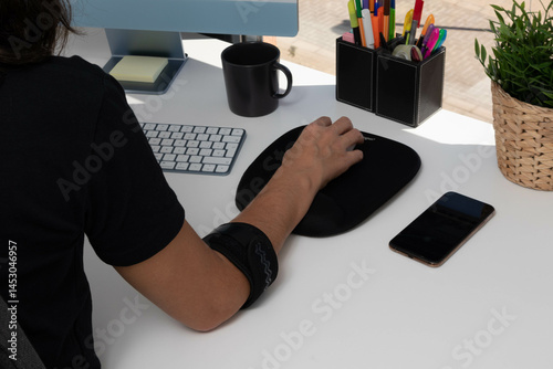 an unrecognizable woman working at computer with tendonitis caused by mouse use, wearing an elbow brace to mitigate the pain