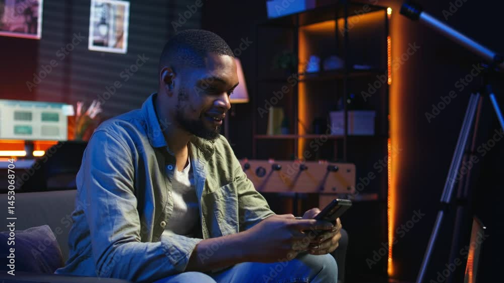 Carefree freelancer chatting with friends over text messages on phone, sitting on the couch and scrolling social media apps. Black guy showcases staying entertained while working remotely. Camera A.