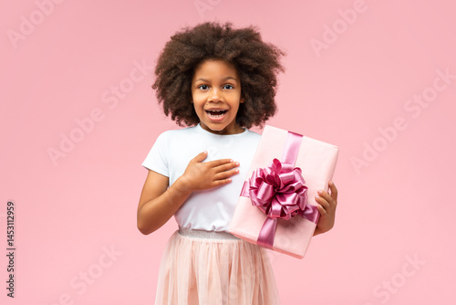Studio portrait of cute joyful African American girl holding decorated gift box and pressing hand to her chest in surprise with excited open mouth face expression, isolated on pink background