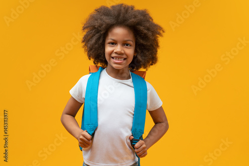 Studio shot of a cute cheerful African American schoolgirl posing over yellow background smiling to the camera and holding straps of her backpack