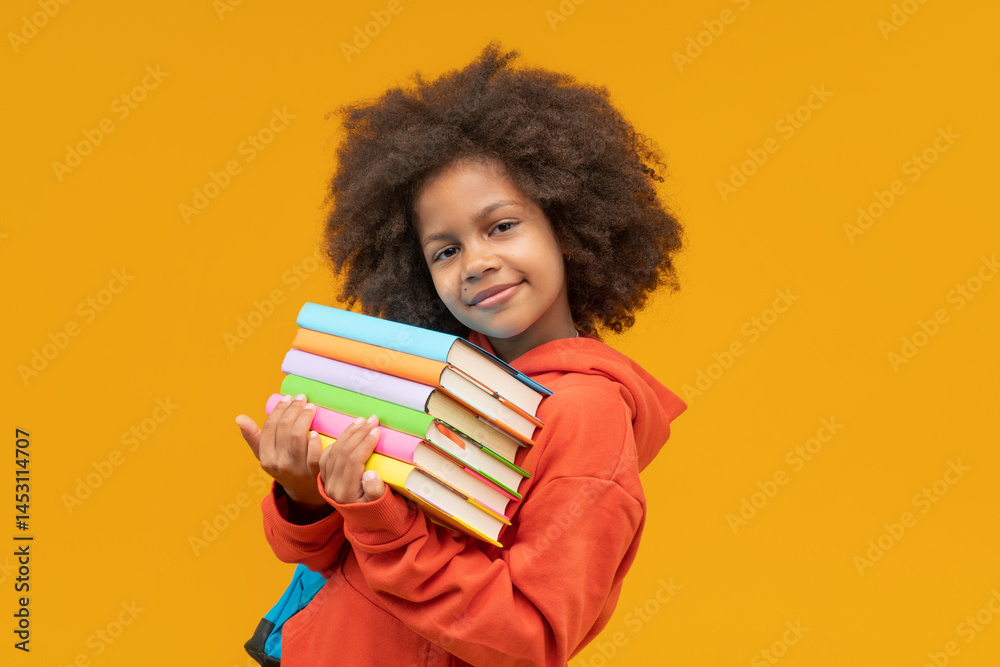 Studio shot of lovely and sweet African American little girl student holding pile of colored books in hands and looking to the camera isolated over yellow background Studio shot of lovely and sweet African American little girl student holding pile of colored books in hands and looking to the camera isolated over yellow background