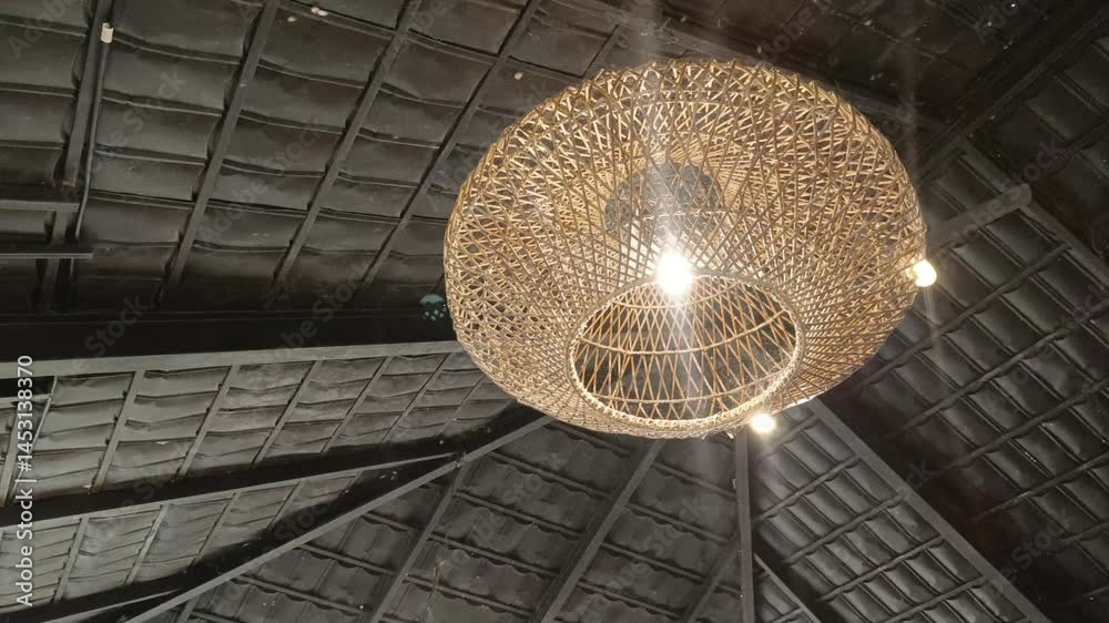 Close up view of a unique wicker pendant lamp hanging against a dark, textured tile ceiling. The fixture adds a touch of craft style to the space.