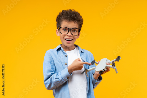 Studio shot of adorable African American schoolboy pointing with index finger at the drone in his hand with excited open mouth face expression, isolated over orange yellow background