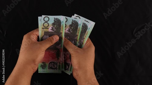 Close-up of a man counting one hundred Australian dollar banknotes with a black background.

