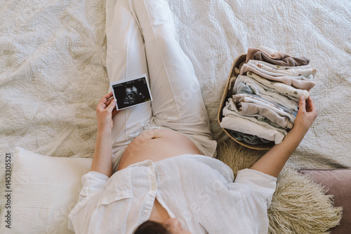 A pregnant woman lies on a bed with an ultrasound image and a basket of baby clothes. The moment of waiting and bonding at home.