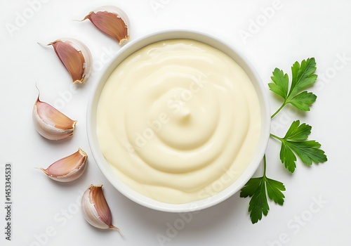 A top view of a bowl of aioli with garlic cloves and parsley sprig on a white surface around the bowl