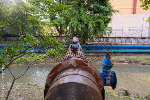  A close-up, eye-level shot focuses on a large, weathered metal pipe running alongside a river. The pipe, showing signs of age and rust, features a complex valve mechanism with blue components.